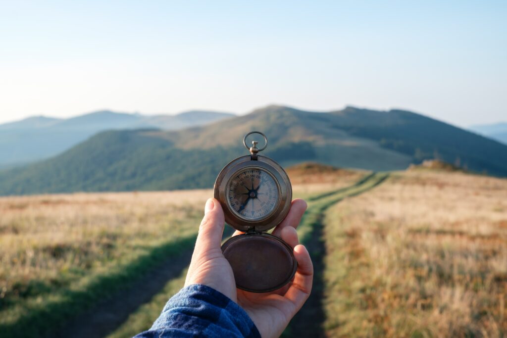 Bild einer Berglandschaft mit großem Feld davor. Mittig hält eine Hand einen Kompass ins Bild.