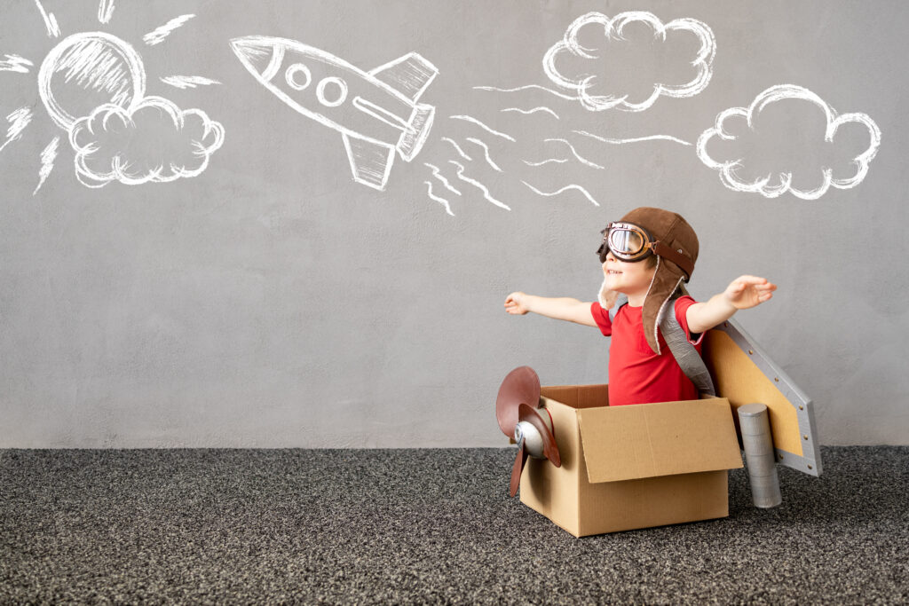 Bild eines kleinen jungen mit Fliegermütze, der in einem zum Flugzeug umgebauten Karton sitzt. Der Junge grinst und streckt die Arme weit ab.
