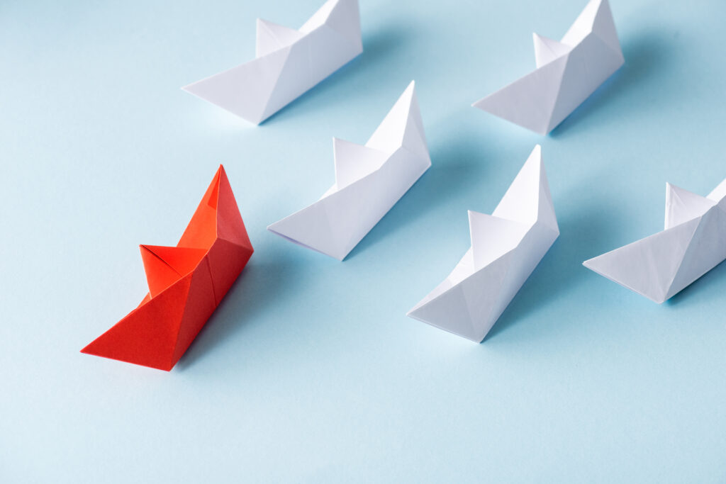 Bild, auf dem sechs Papierschiffchen abgebildet sind. Ein rotes führt fünf weiße an.