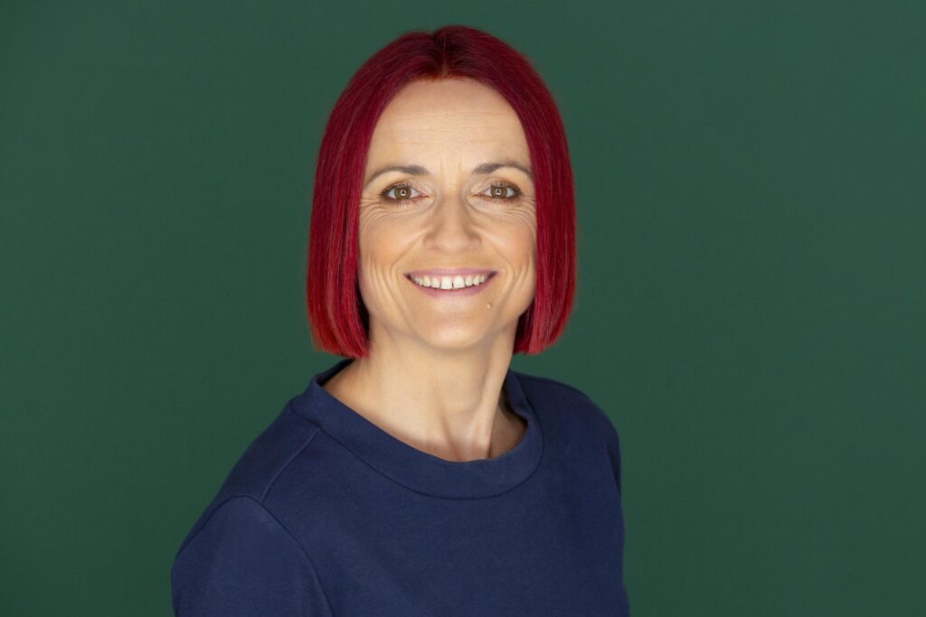 Porträtfoto von Vicky Benub, Geschäftsführerin der zeitraum Ravensburg Praxis für Boeger und Massage. Sie steht vor einem grünen Hintergrund, hat kurze rote Haare, trägt einen blauen Pullover und lächelt freundlich.