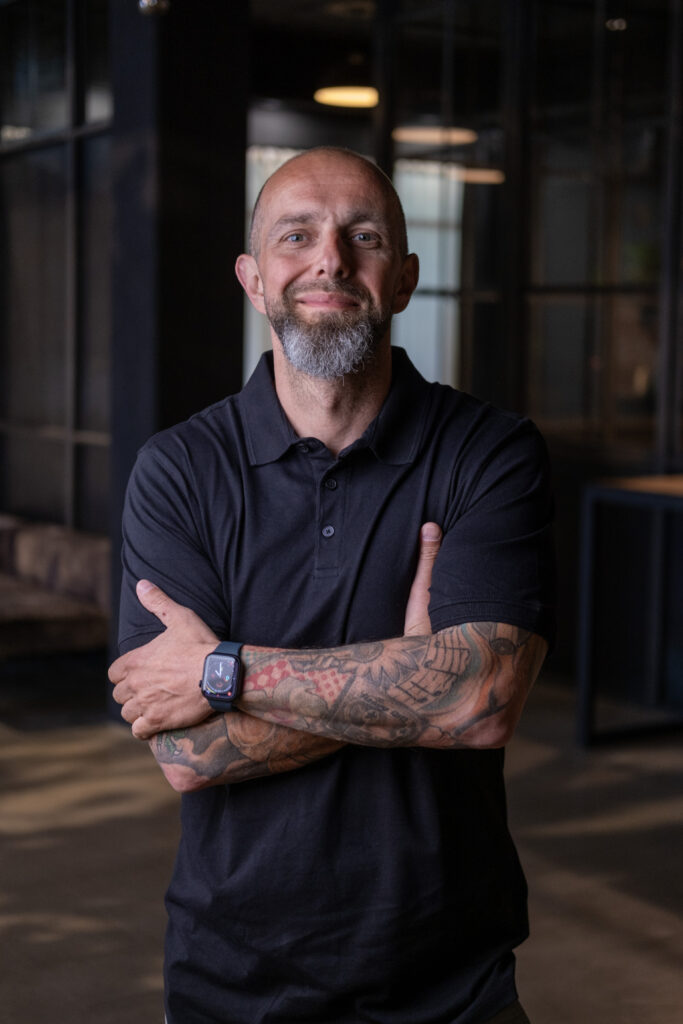 Foto von Mike Krenzien von Digital Apes. Er trägt ein schwarzes, kurzärmeliges Hemd, hat die Arme lässig verschränkt und lächelt freundlich.